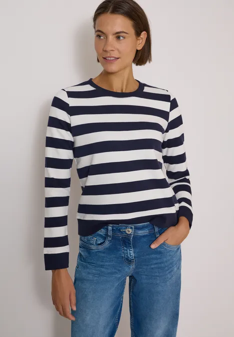 Frau mit dunkelblau-weiß gestreiftem Pullover, blauer Jeans und kurzen braunen Haaren.