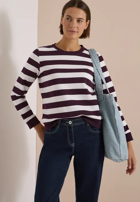 Frau in burgund-weiß gestreiftem Langarmshirt, Jeans und mit hellblauer Jeans-Tasche vor hellem Wandhintergrund.