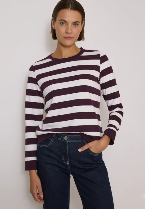 Frau in weiß-dunkel gestreiftem Pullover und Jeans vor hellem Hintergrund.