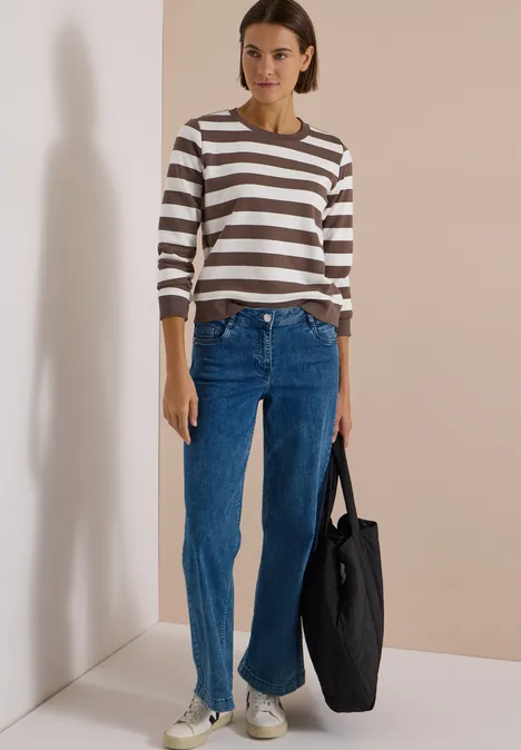 Frau in gestreiftem Pullover, Jeans und schwarzer Tasche steht vor beigem Hintergrund.