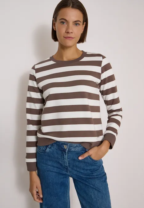 Frau in braun-weiß gestreiftem Langarmshirt und Jeans steht vor hellem Hintergrund.