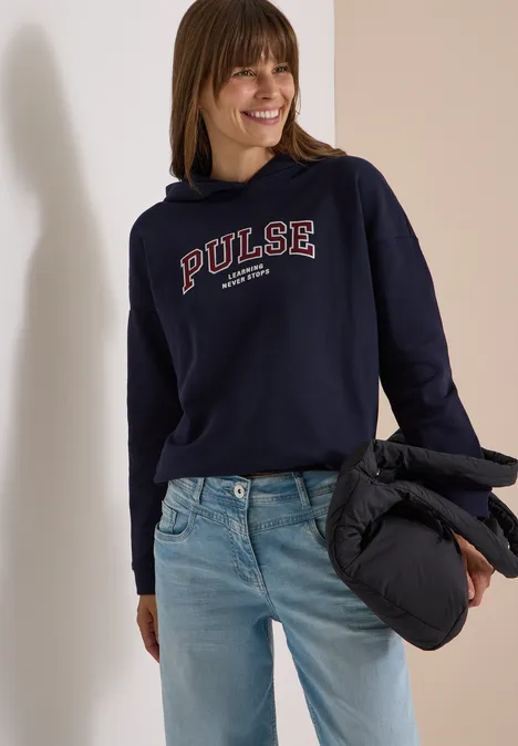 Femme souriante en sweat à capuche bleu marine « PULSE », jean clair et veste matelassée noire à la main.