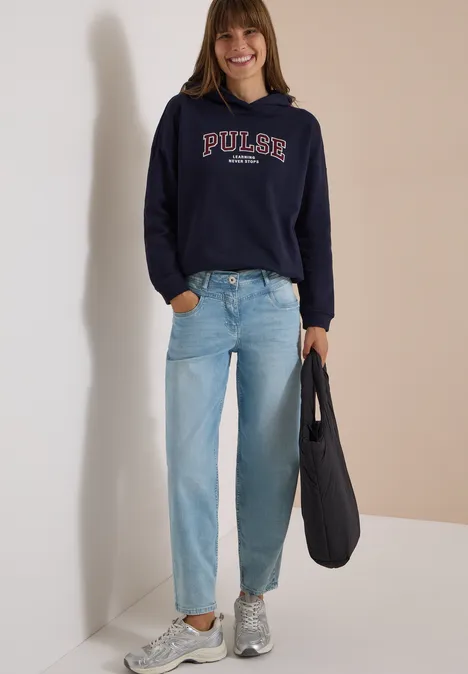 Une femme souriante porte un sweat bleu marine 'PULSE', un jean clair, des baskets grises et un sac noir.