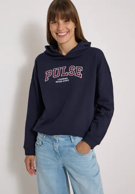 Femme en sweat à capuche bleu marine 'PULSE' et jean bleu, souriante sur fond clair.