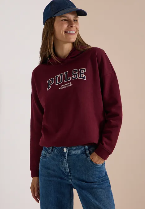 Femme souriante en sweat à capuche bordeaux 'PULSE', jean bleu et casquette de baseball.