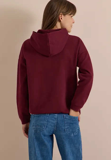 Vue de dos d'une femme en jean et sweat à capuche bordeaux sur fond clair.