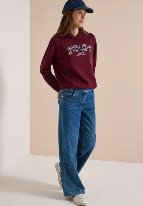 Femme en tenue décontractée avec sweat bordeaux, jean large bleu, baskets et casquette.