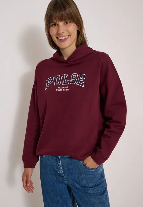 Jeune femme portant un sweat à capuche bordeaux 'PULSE' et un jean bleu sourit devant un fond clair.