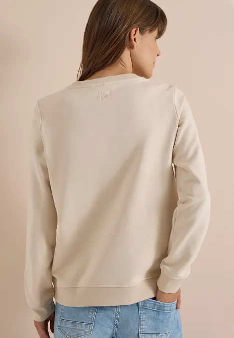 Une femme aux cheveux bruns porte un sweat-shirt beige clair et un jean bleu, vue de dos.