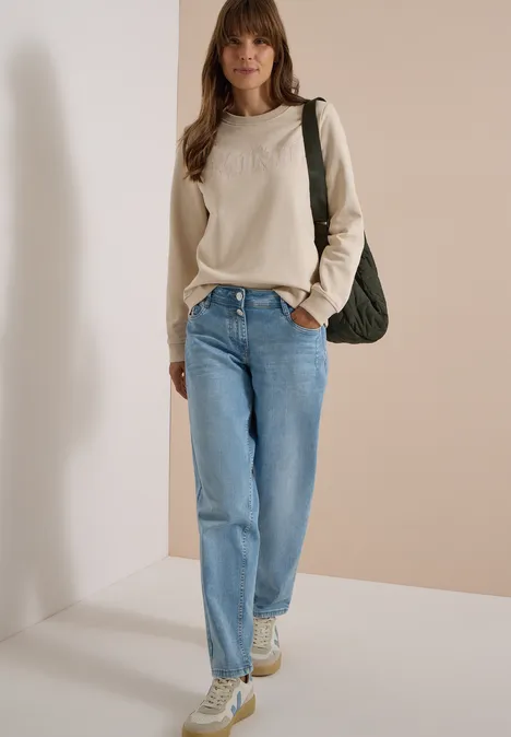 Femme en pull beige, jean bleu et baskets, portant un sac vert devant un mur beige clair.