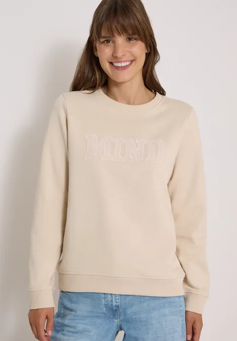 Femme en sweat-shirt beige 'MIND' et jean bleu, souriante sur fond clair.