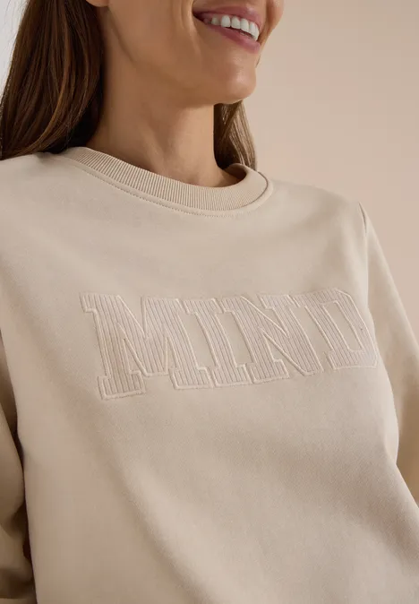 Femme de profil porte un sweat écru brodé «MIND» et sourit légèrement.