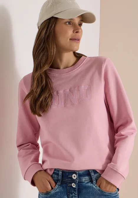 Femme porte un sweat rose, un jean bleu et une casquette beige.