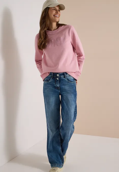 Jeune femme souriante en pull rose, jeans bleus et casquette blanche appuyée contre un mur.