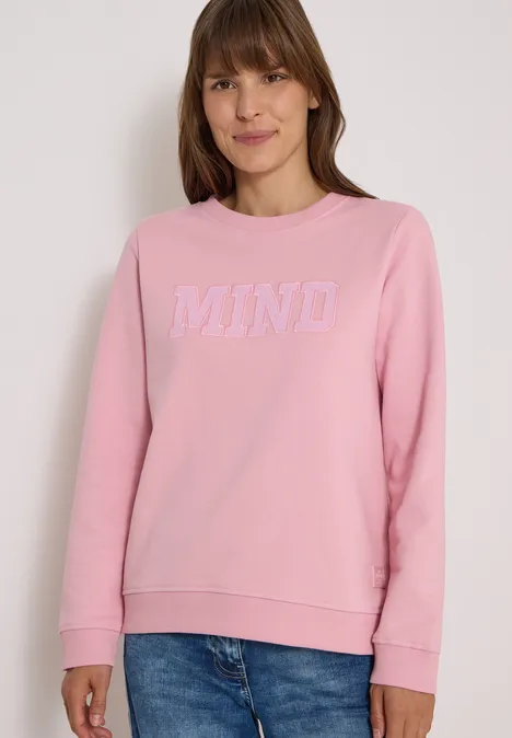 Femme portant un sweat-shirt rose avec l’inscription blanche "MIND", souriante.