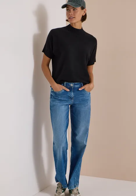 Femme en t-shirt noir, jean bleu et casquette, adossée à un mur clair.