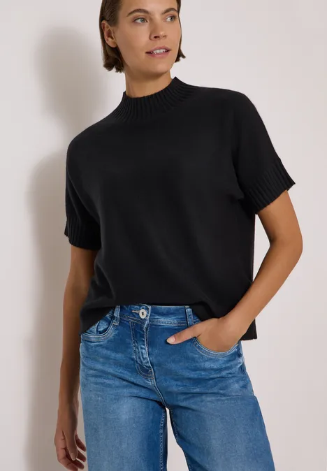 Femme portant un pull noir à manches courtes et un jean bleu sur fond clair.