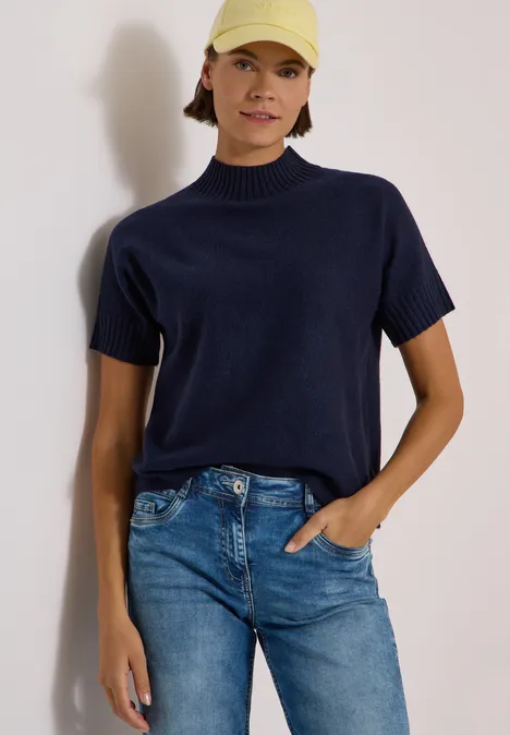 Femme portant un pull court bleu marine, jean et casquette jaune clair sur fond clair.