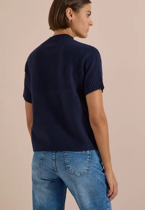 Femme de dos portant un pull marine à manches courtes et un jean bleu sur fond beige.