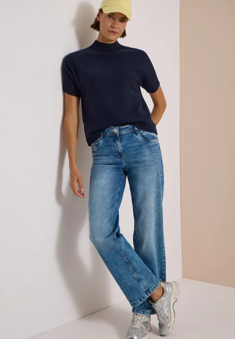 Femme adossée à un mur clair, polo bleu marine, jean droit, baskets blanches et casquette jaune.