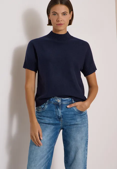 Femme portant un pull à col montant bleu à manches courtes et un jean droit bleu, fond blanc.