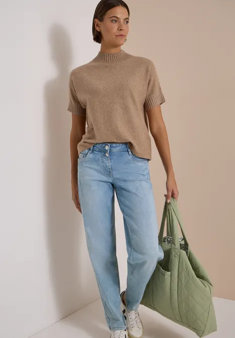 Femme appuyée contre un mur, pull beige à col montant, jean bleu clair, baskets blanches et sac matelassé vert.
