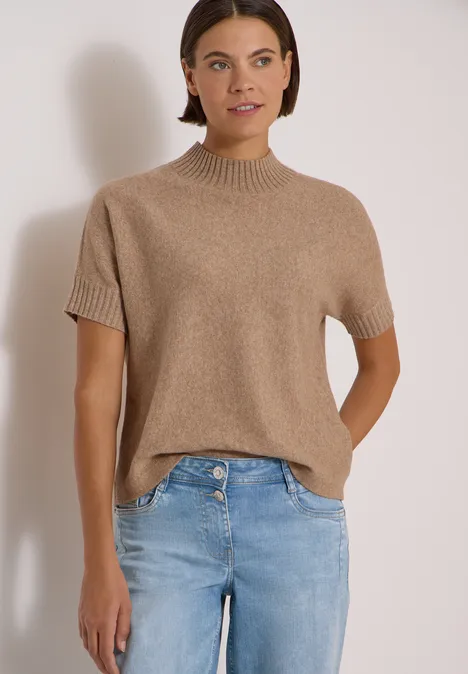 Une femme porte un pull en maille beige à manches courtes et un jean bleu devant un fond clair.
