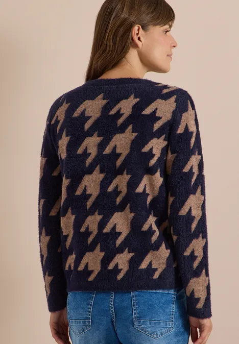 Vrouw van achteren in een marine-beige wollen trui met houndstoothpatroon en blauwe jeans, lichte achtergrond.