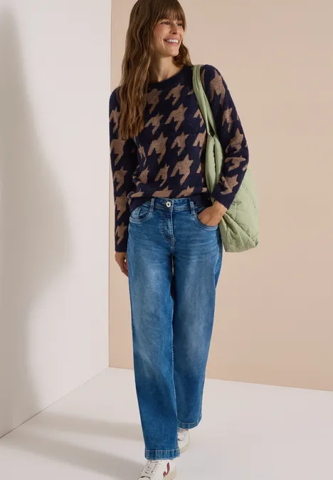 Vrouw glimlacht in pied-de-poule trui, wijde jeans en groene schoudertas, voor neutrale achtergrond.