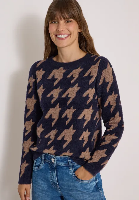Vrouw draagt marineblauwe trui met beige pied-de-poule patroon en spijkerbroek, lacht