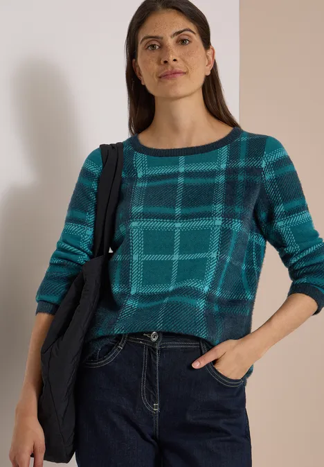 Frau in kariertem Türkis-Pullover und Jeans, lässige Schultertasche, Hand in Tasche.