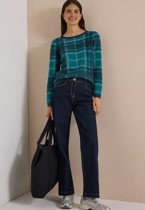 Frau in kariertem grünen Pullover, weiten dunkelblauen Jeans und großer Tasche.