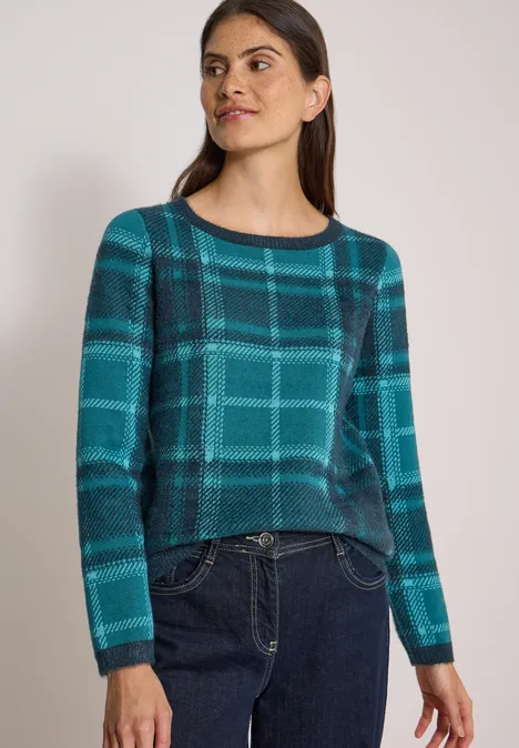 Frau trägt langärmligen Pullover mit türkis-blauem Karomuster und Jeans.