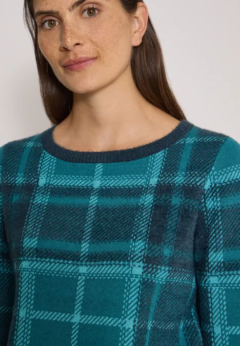 Frau mit dunklem Haar und Sommersprossen trägt einen karierten Teal-Schwarz-Pullover mit Rundhalsausschnitt.