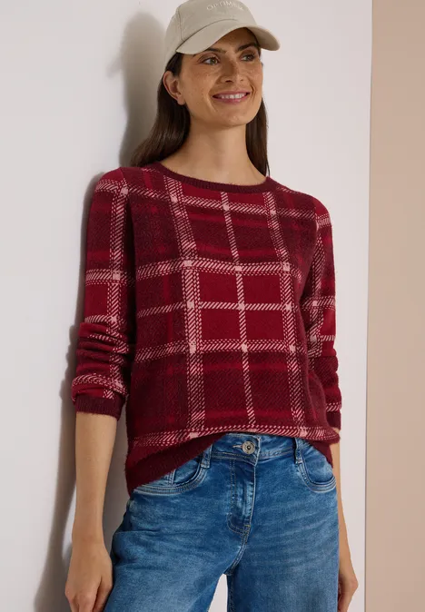 Langarm Pullover mit Karomuster red wine Frau trägt roten karierten Pullover, blaue Jeans und Cap, steht vor hellem Hintergrund.