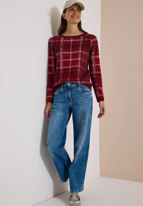 Langarm Pullover mit Karomuster red wine Frau in rotem Karopullover, blauen Jeans, grünen Sneakern und Cap vor neutralem Hintergrund.