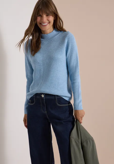 Pull texturé avec des boutons décoratifs easy blue melange Femme souriante en pull bleu clair et jean foncé, tenant une veste verte devant un fond neutre.
