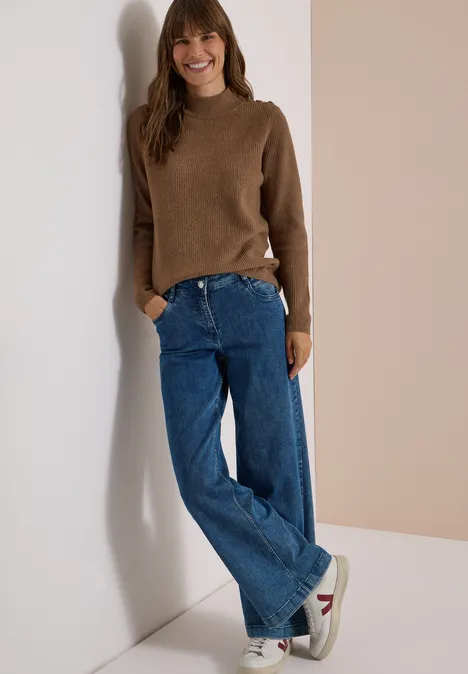 Pull texturé avec des boutons dark toffee brown melange Une femme souriante en pull marron, jean large bleu et baskets appuyée contre un mur.