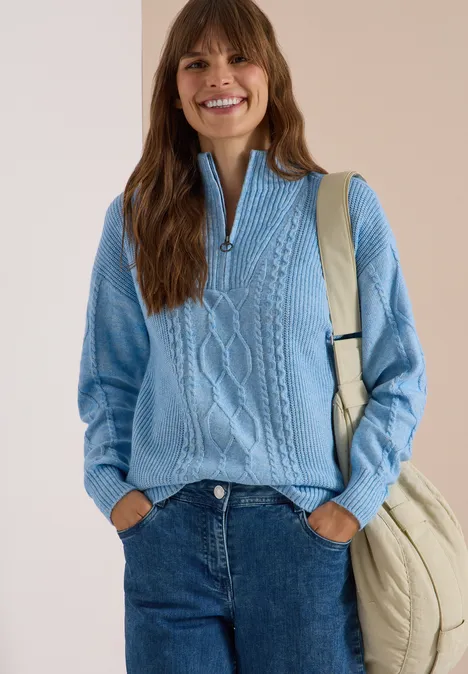 Femme souriante en pull torsadé bleu à demi-fermeture, jean et sac beige sur l'épaule, fond lumineux.