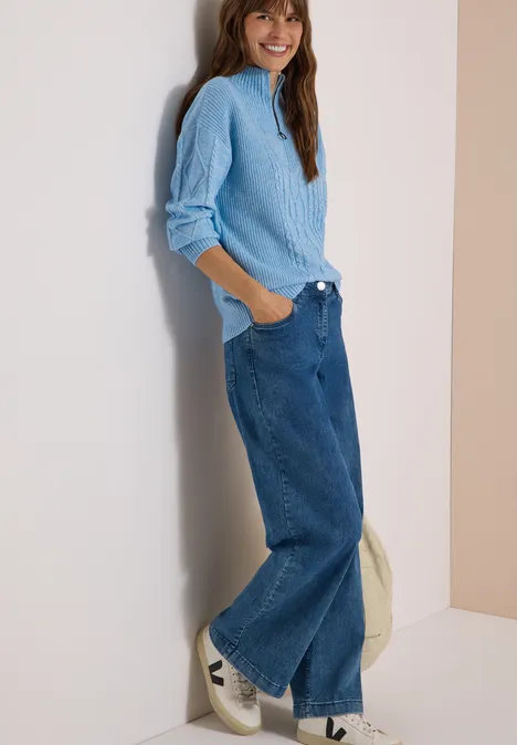 Femme adossée à un mur, pull bleu clair, jean large et baskets blanches, sourire décontracté.