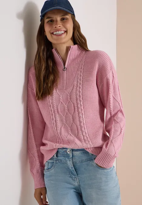 Une femme souriante portant un pull rose torsadé à col zippé, un jean et une casquette bleu marine.