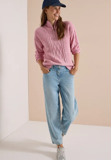 Femme en pull rose, jean ample clair et baskets, posant devant un mur neutre.