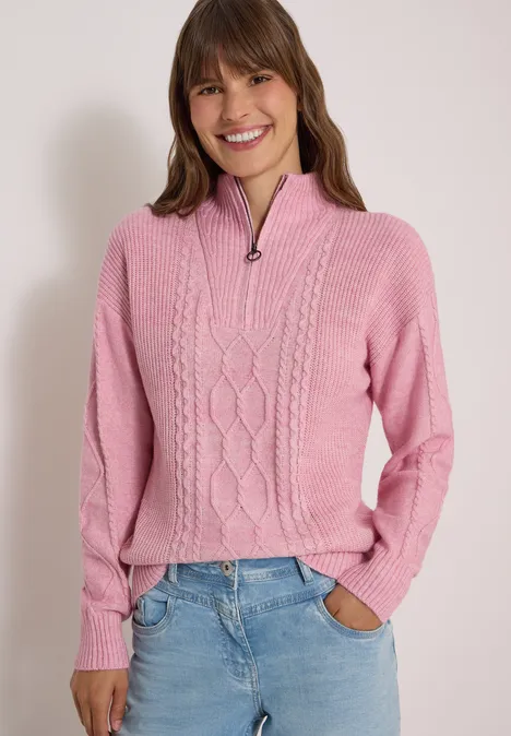 Une femme souriante en pull torsadé rose à demi-fermeture éclair et jean bleu.