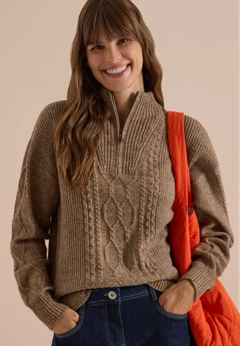 Une femme souriante portant un pull en maille brune torsadée à demi-fermeture éclair et un sac orange.