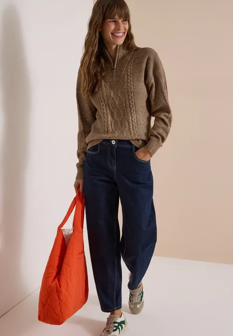 Femme en pull cable marron, jeans bleus et baskets vertes, portant un sac rouge