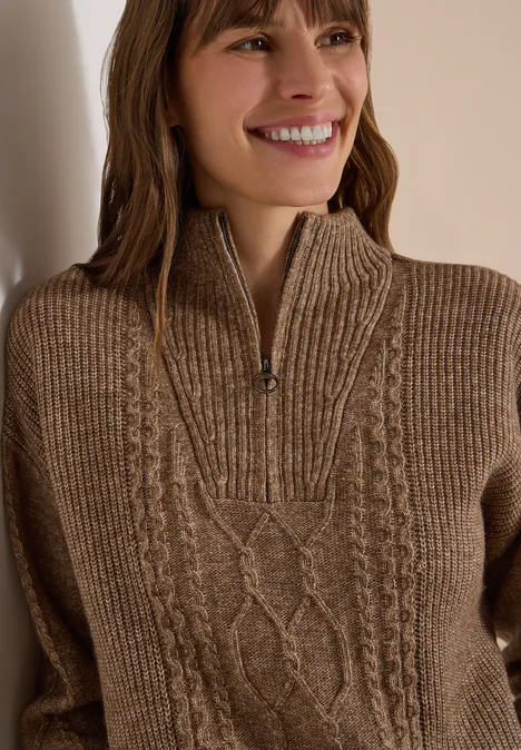 Femme souriante portant un pull marron à grosse maille, motif torsadé et col zippé mi-haut.