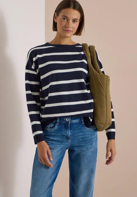 Frau in gestreiftem Pullover, Jeans und grünem Steppbeutel vor hellem Hintergrund.