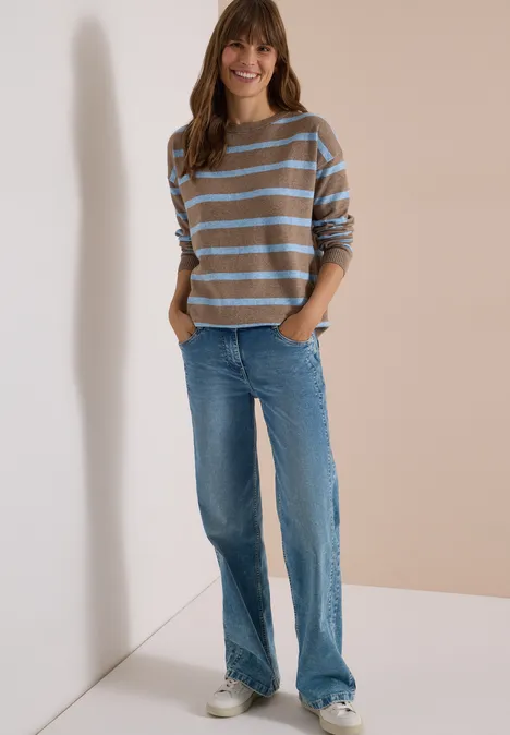 Eine junge Frau in gestreiftem Pullover, blauen Jeans und Sneakers steht lächelnd vor neutralem Hintergrund.