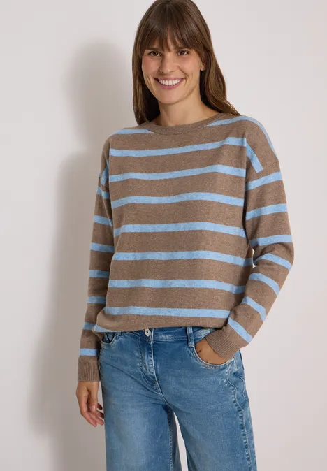 Frau trägt gestreiften Pullover in Braun und Hellblau sowie Jeans, lächelt und hat eine Hand in der Hosentasche.