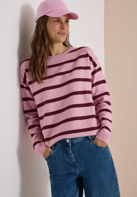 Pull à col bateau et rayures mellow rose melange Femme en pull rayé rose et violet, jean et casquette rose, appuyée détendue contre un mur.